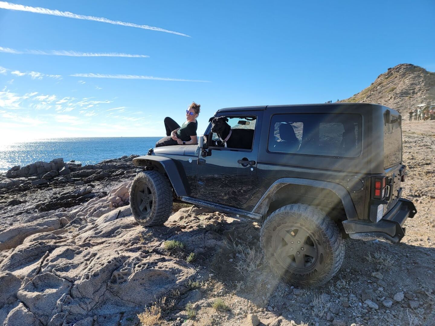 A woman sitting on the hood of a Jeep on a rocky beach, with a black dog looking out the window of the driver's seat.