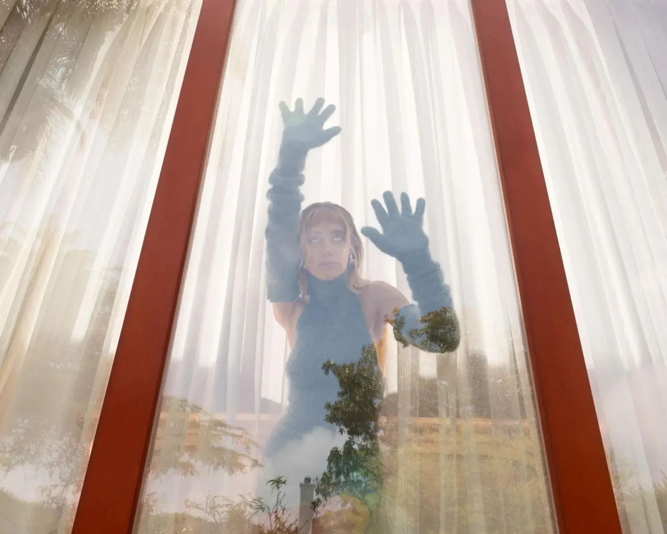 Still of Longlegs’ star Maika Monroe with her gloved hands leaning against a window.